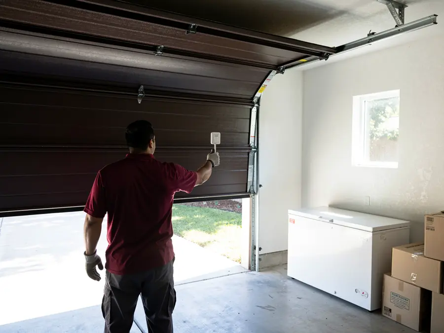 A technician in a red shirt is holding a white device near a brown garage door.